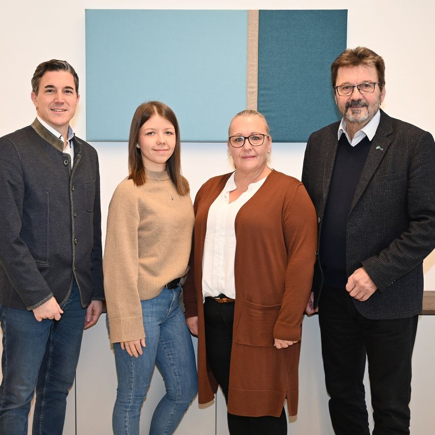 Vier Personen stehen nebeneinander vor einer blau-weißen Wand. Sie alle lächeln und scheinen für ein Foto zu posieren.