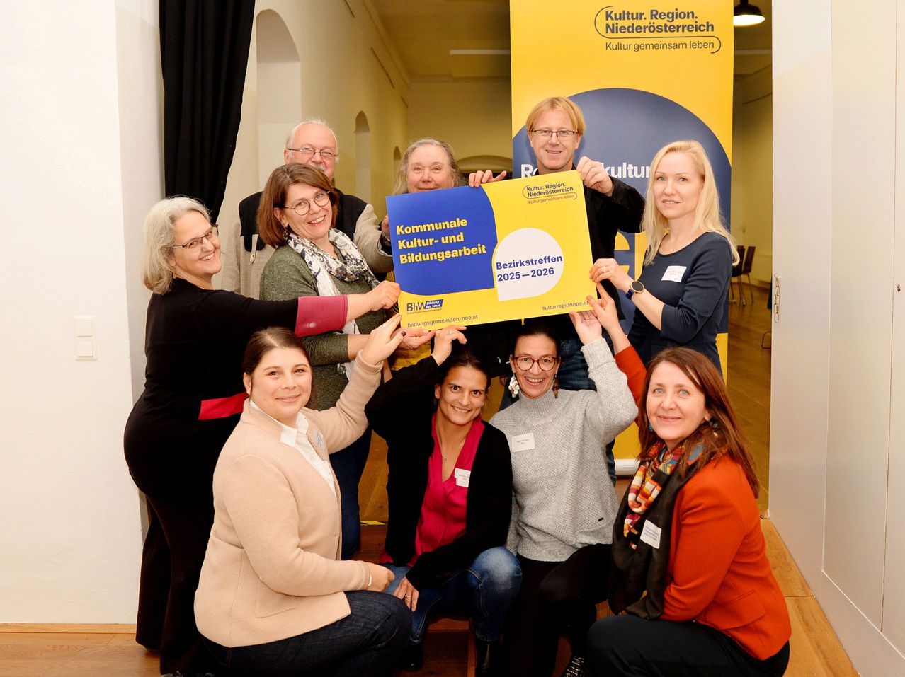 Eine Gruppe von Menschen posiert für ein Foto mit einem Banner, das 'Kommunale Kultur- und Bildungsarbeit Bezirkstreffen 2025-2026' in einem Raum liest.