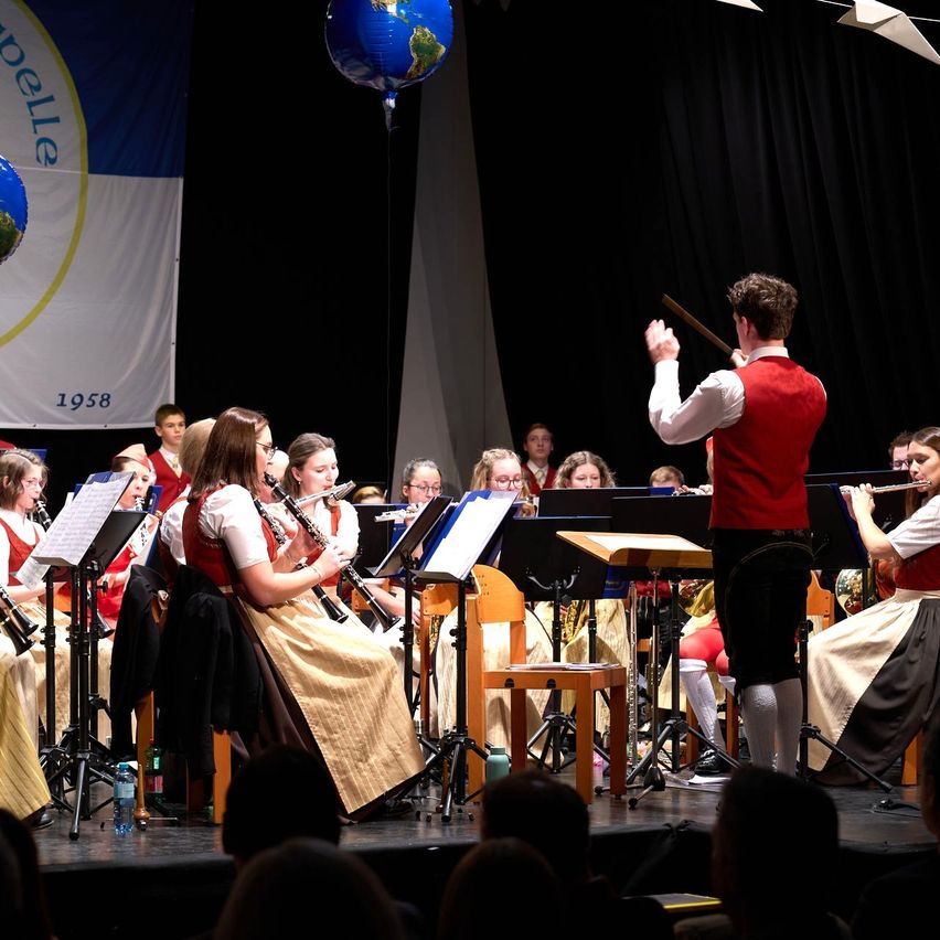 Ein musikalischer Auftritt mit einem Dirigenten, der eine Gruppe von Musikern anführt, die verschiedene Instrumente auf der Bühne vor einem Publikum spielen.