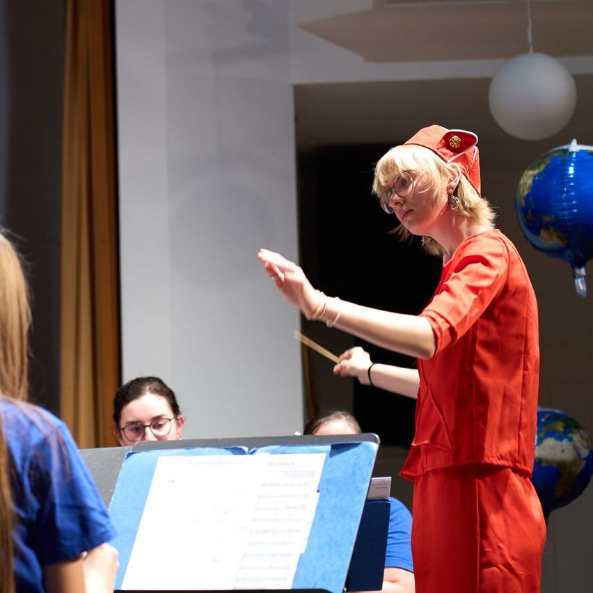 Eine Frau in einem roten Outfit steht vor einem Notenständer und dirigiert mit einem Taktstock. Hinter ihr sitzt eine Person, die Musik liest. Zwei weitere Personen sind im Hintergrund, und eine Erdkugel hängt von der Decke.