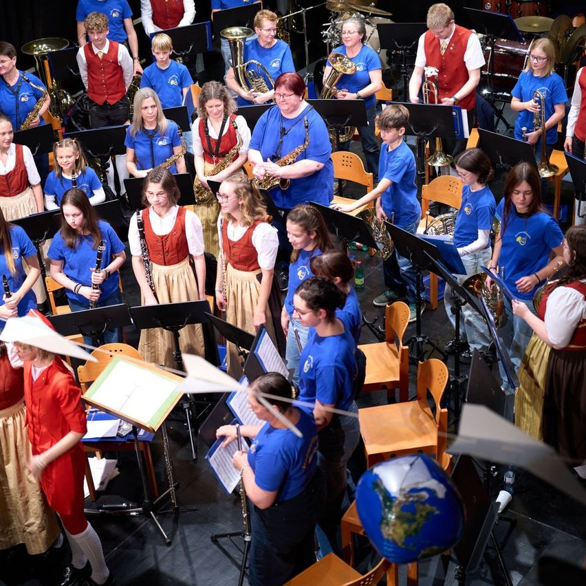 Ein Schulorchester probet auf der Bühne, wobei die Mitglieder um verschiedene Musikinstrumente herum stehen und sitzen. Einige spielen Trompeten, während andere Saxophone spielen. Der Raum hat eine Bühne, Stühle und einen schwarzen Boden.