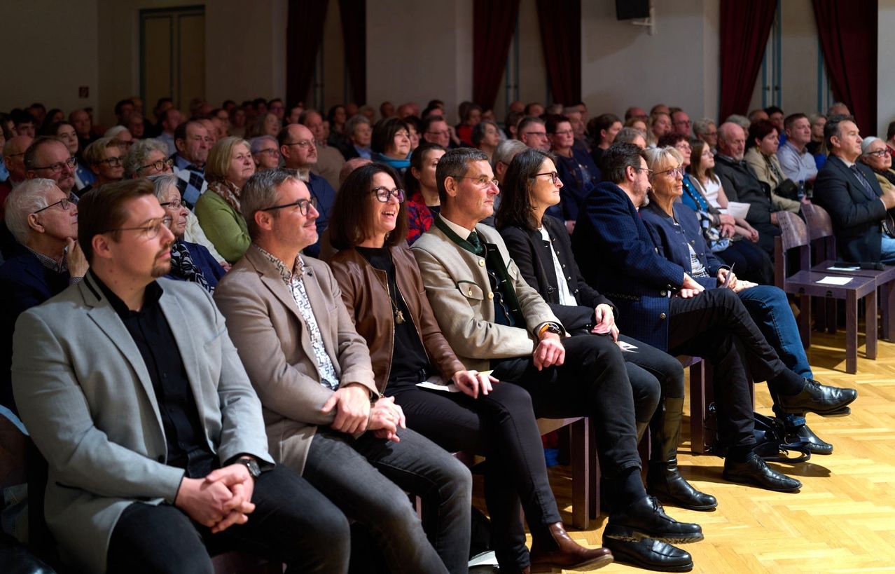 Eine Menschenmenge sitzt auf Bänken in einem Raum. Einige tragen Brillen, und einige haben Schals. Sie lauschen aufmerksam. Dahinter sitzen weitere Menschen, und einige stehen.