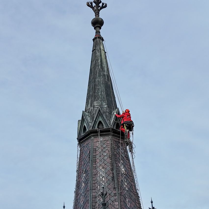 Eine Person in einem roten Sicherheitsanzug klettert mit Seilen und Werkzeugen einen Kirchturm. Der Turm besteht aus Ziegelsteinen und hat ein spitzes Dach. Der Himmel ist klar.