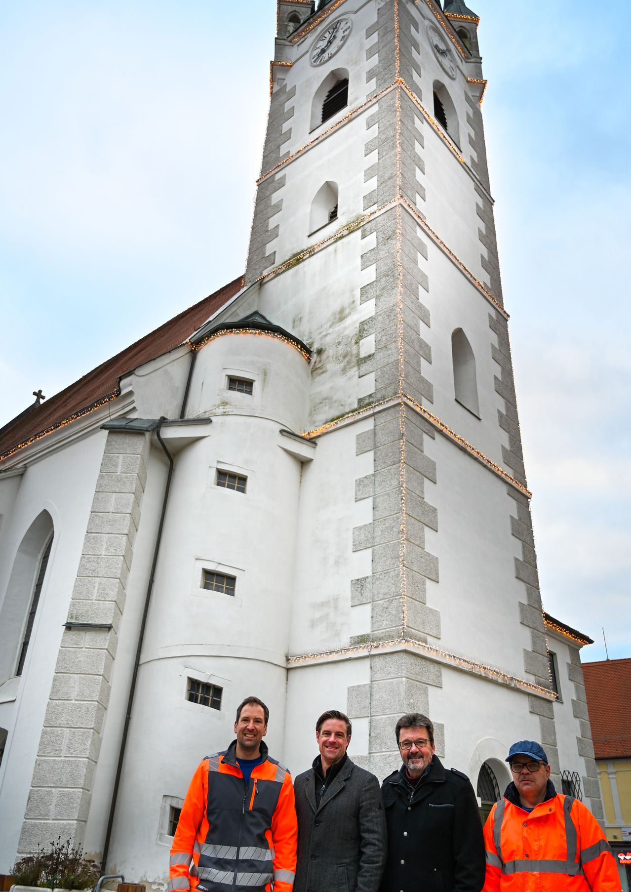 Drei Männer stehen vor einer Kirche mit einem hohen Turm und dekorativen Lichtern. Sie lächeln.