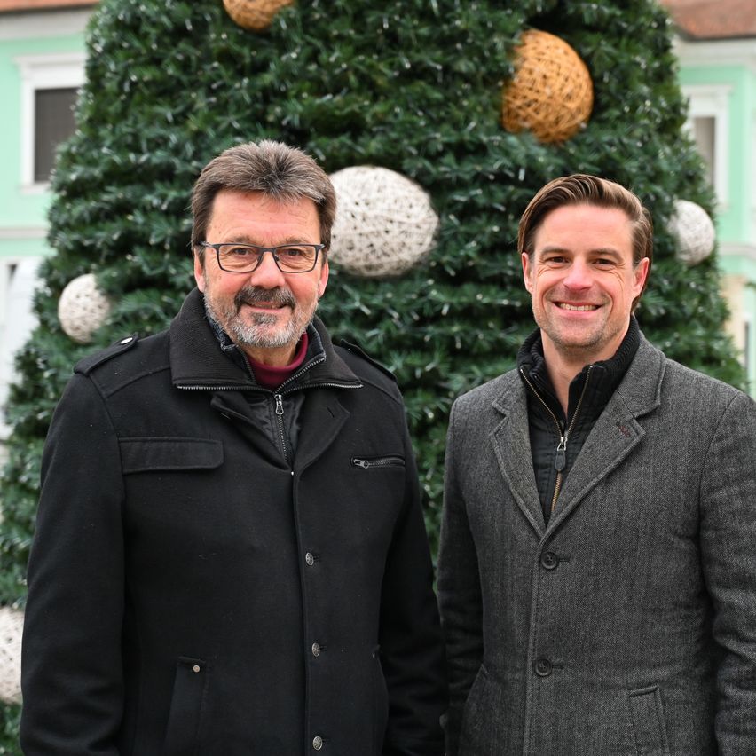 Zwei Männer stehen Seite an Seite vor einem Weihnachtsbaum. Einer trägt einen schwarzen Mantel, der andere einen grauen. Beide lächeln.