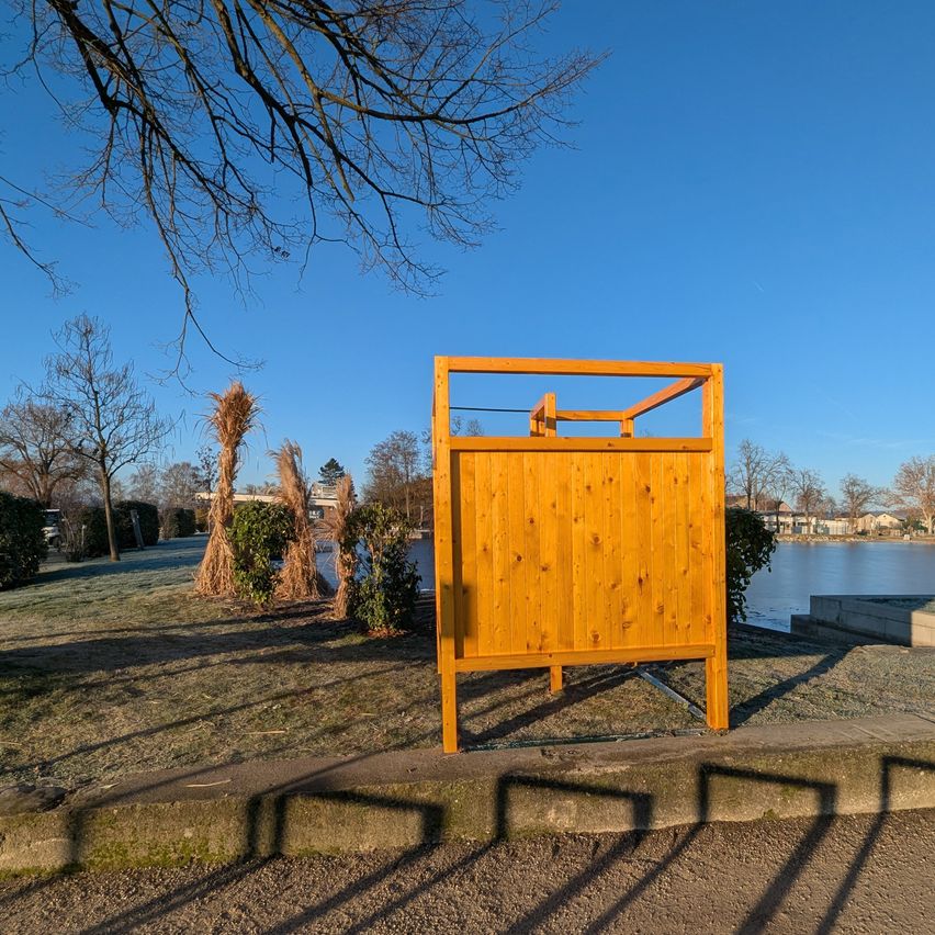 Eine Holzkonstruktion steht auf einer Rasenfläche in der Nähe eines Sees. Der Himmel ist klar und Schatten fallen auf den Boden.