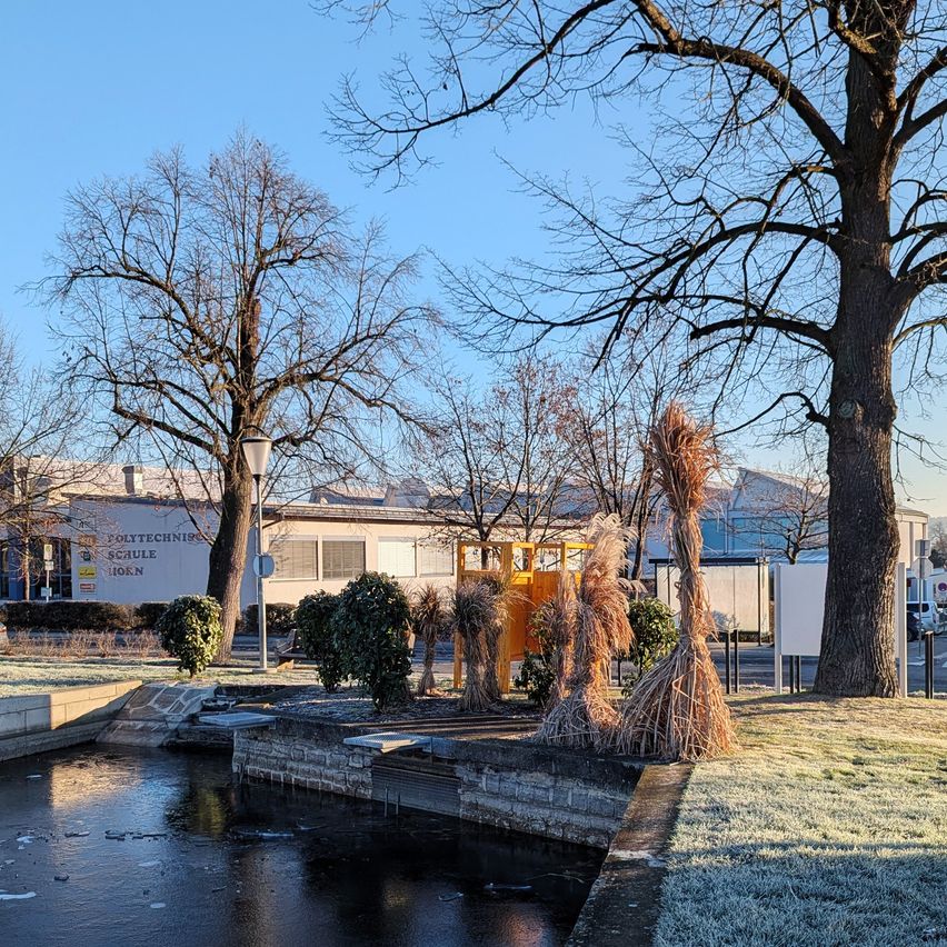 Eine friedliche Szene eines teilweise gefrorenen Teichs mit einem Gebäude namens Polytechnik Schule im Hintergrund unter einem klaren blauen Himmel.