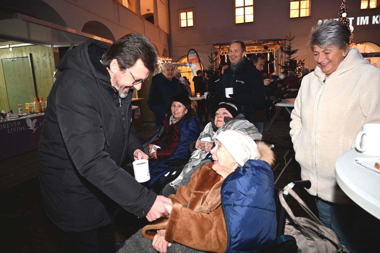 Ein Mann reicht einer sitzenden älteren Frau eine Tasse, beide lächeln, mit mehreren anderen Personen und einem Gebäude im Hintergrund.