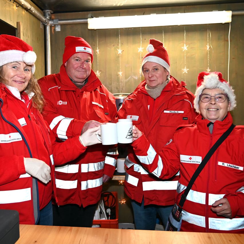 Fünf Personen in roten Jacken und Weihnachtsmützen stehen zusammen und halten weiße Tassen. Im Hintergrund befindet sich eine mit Weihnachtsdekorationen und einer Lampe verzierte Wand. Ein roter Aufbewahrungskasten steht auf dem Boden.