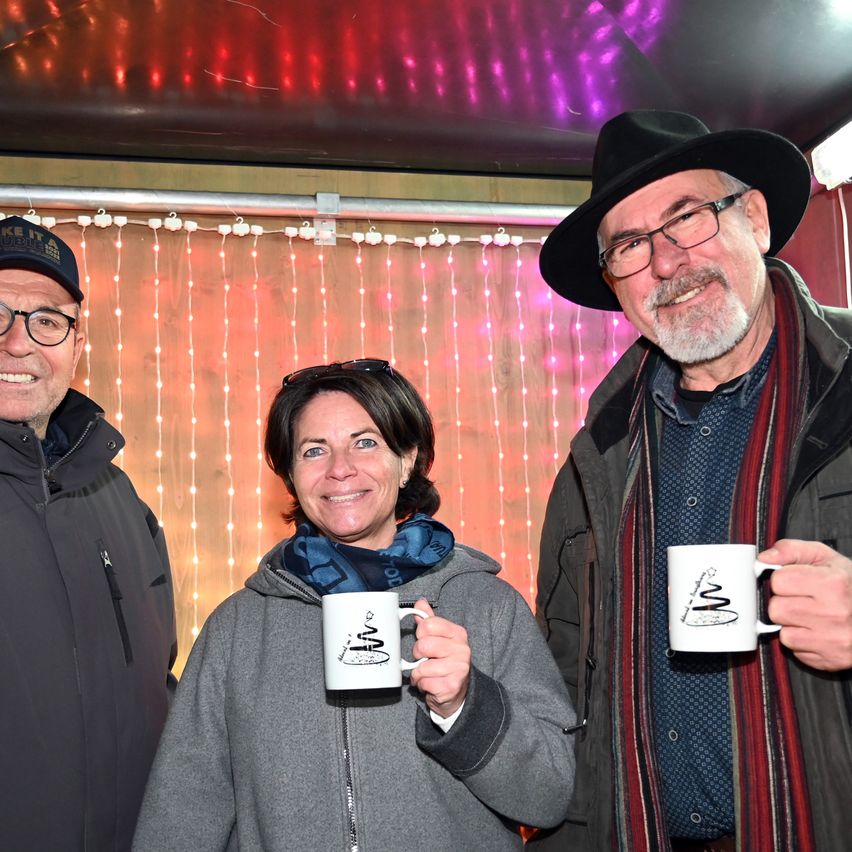 Drei Personen stehen unter einem Vordach mit dekorativen Lichtern. Ein Mann links trägt einen Hut und eine Brille, eine Frau in der Mitte lächelt und hält eine Kaffeetasse, und ein Mann rechts hält ebenfalls eine Kaffeetasse.