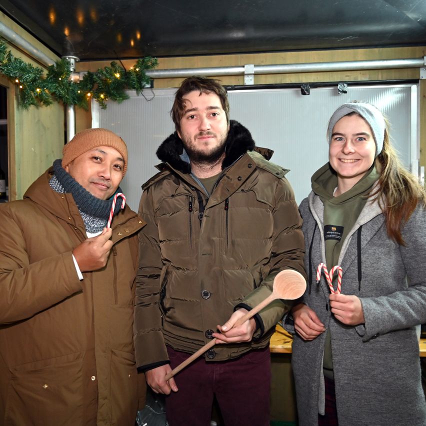 Drei Menschen, warm gekleidet, stehen in einem weihnachtlich dekorierten Zelt. Ein Mann und eine Frau lächeln und halten Zuckerstangen und einen Holzlöffel. Hinter ihnen schmücken weihnachtliche Dekorationen die Wände.