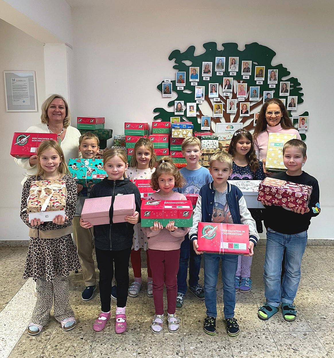 Eine Gruppe von Kindern und Erwachsenen posiert mit Geschenkboxen für ein Foto, vor einem großen Baum mit Fotoschmuck.