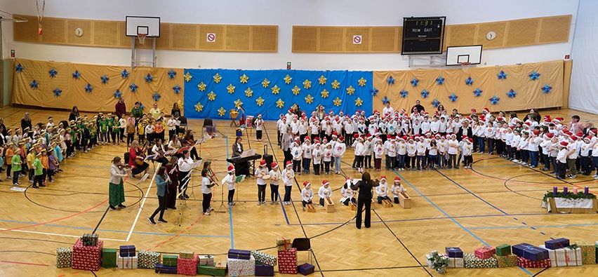 Eine Gruppe von Menschen in einer Turnhalle mit Weihnachtsthema, einige mit Weihnachtsmützen, stehen um einen Kreis von Geschenken und einer zentralen Versammlung. Die Turnhalle hat blaue Linien auf dem Boden und Dekorationen an den Wänden.