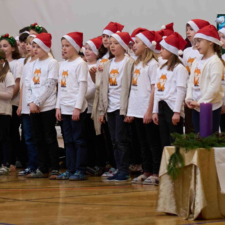 Eine Gruppe von Kindern, alle mit Weihnachtsmützen, steht in einer Reihe. Sie singen wahrscheinlich ein Weihnachtslied. Einige tragen Turnschuhe, andere Sandalen. Hinter ihnen ist eine weiße Wand mit Weihnachtskränzen geschmückt. Ein Tisch mit einer lila Kerze steht rechts.