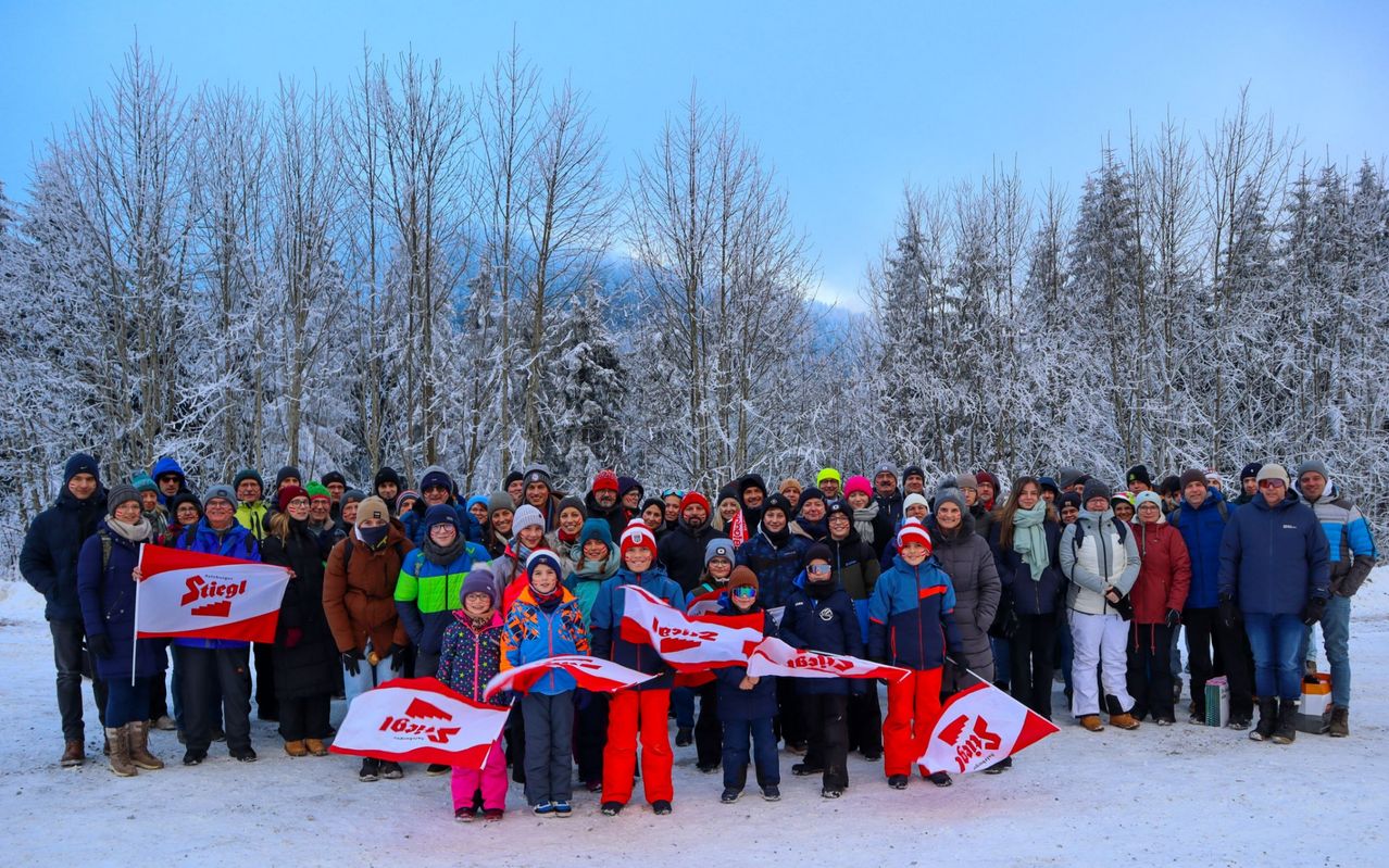 Eine Gruppe von Menschen versammelt sich in einer verschneiten Landschaft, um ein Foto zu machen. Sie tragen Winterkleidung, einige mit Mützen und Brillen. Mehrere Fahnen mit dem Wort 'Ski' sind sichtbar.