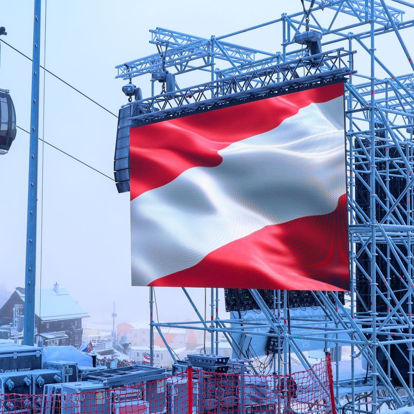 Eine große österreichische Flagge ist auf einer Metallkonstruktion in einer verschneiten Landschaft zu sehen, mit einer Gondel und Gebäuden im Hintergrund.