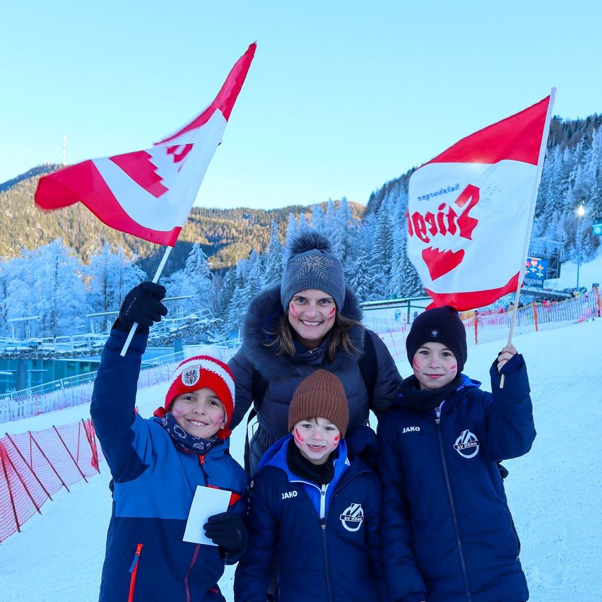 Eine Frau und drei Kinder stehen in einem verschneiten Bereich, halten Fahnen und lächeln. Sie sind alle warm gekleidet. Dahinter befindet sich ein schneebedeckter Berg und ein Skigebiet.