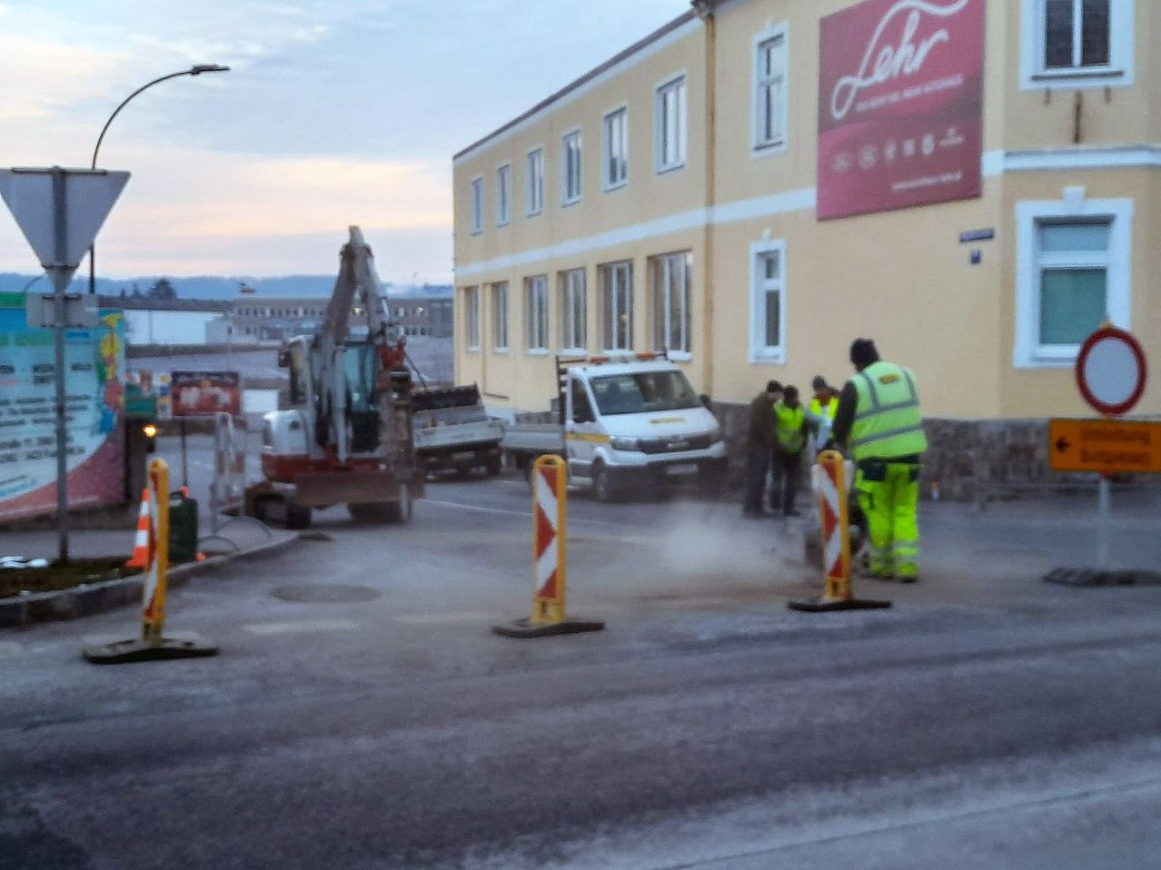 Eine Baucrew arbeitet auf einer Straße, mit einem Baufahrzeug und Arbeitern, die in der Nähe eines Gebäudes stehen, an dem ein Schild mit der Aufschrift Lehr angebracht ist.