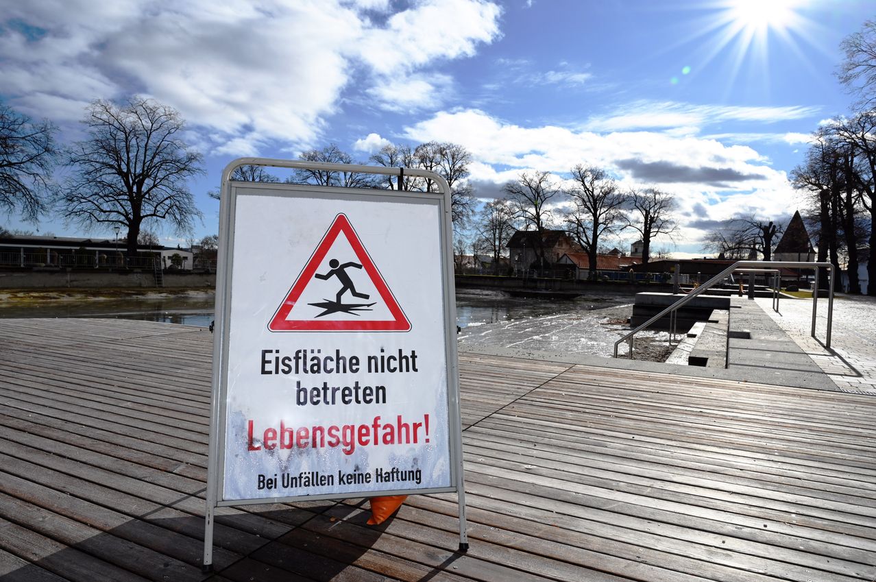 Ein weißes Schild mit einem roten Dreieck und einer schwarzen Figur warnt vor Eisgefahren. Auf dem Schild steht 'Eisflächen nicht betreten, Lebensgefahr!'. Es befindet sich auf einer Holzterrasse.