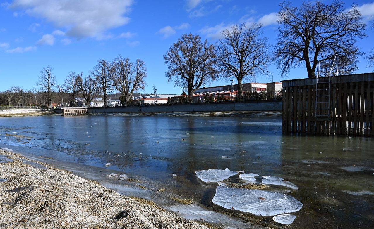 Ein gefrorener Fluss mit mehreren Eisbrocken am Ufer, kahlen Bäumen und einem Gebäude im Hintergrund unter einem blauen Himmel mit einigen Wolken.