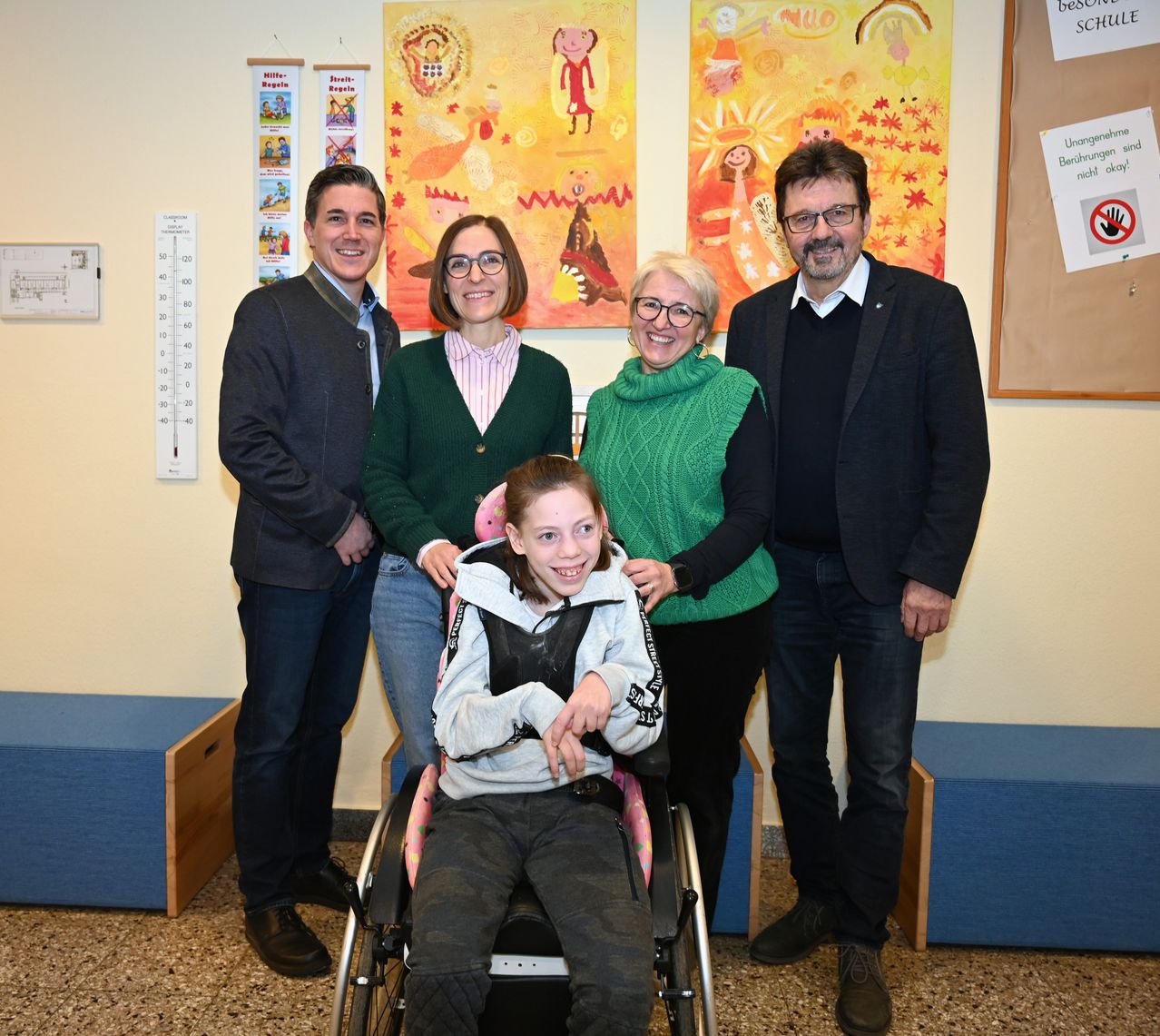 Fünf Personen posieren für ein Foto in einem Raum mit farbenfrohen Gemälden und einem Thermometer an der Wand. Eine junge Mädchen im Rollstuhl steht im Mittelpunkt.