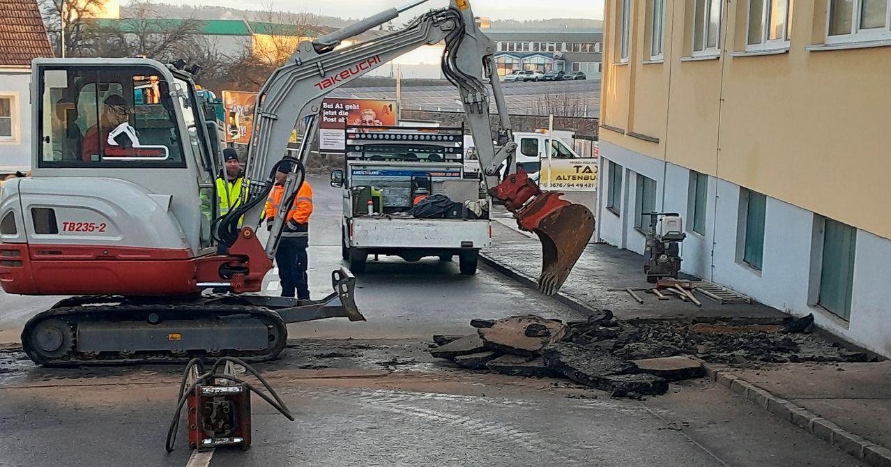 Ein Baufahrzeug mit Takeuchi-Marke ist auf einer Straße geparkt. Ein Arbeiter in Schutzkleidung steht in der Nähe. Haufen von Schutt sind auf der Straße verstreut. Ein Gebäude befindet sich rechts.