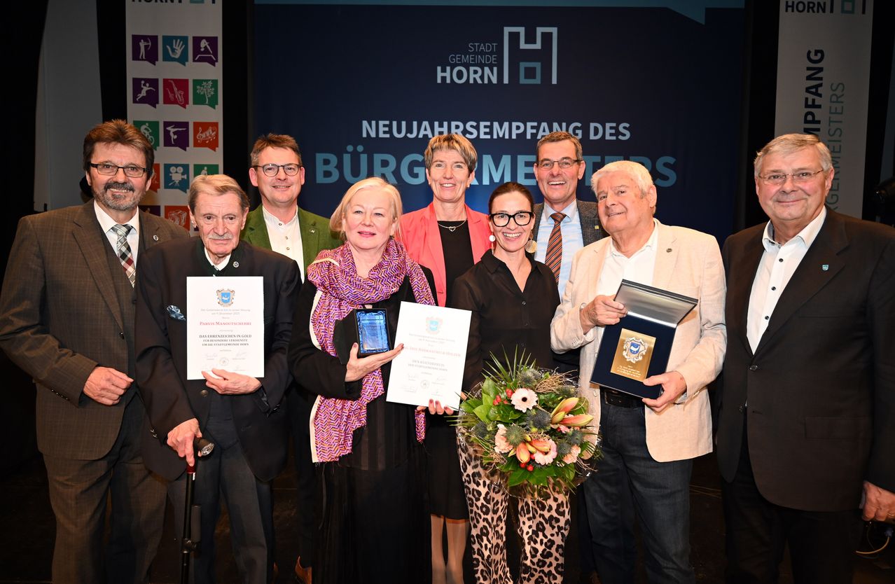 Eine Gruppe von formell gekleideten Personen, die Auszeichnungen und Urkunden in den Händen halten, steht gemeinsam auf einer Bühne mit einem Hintergrund, der Stadtlogos und die Worte 'Neujahrsempfang des Burgermeisters' zeigt. Eine Frau in der Mitte hält einen Blumenstrauß.