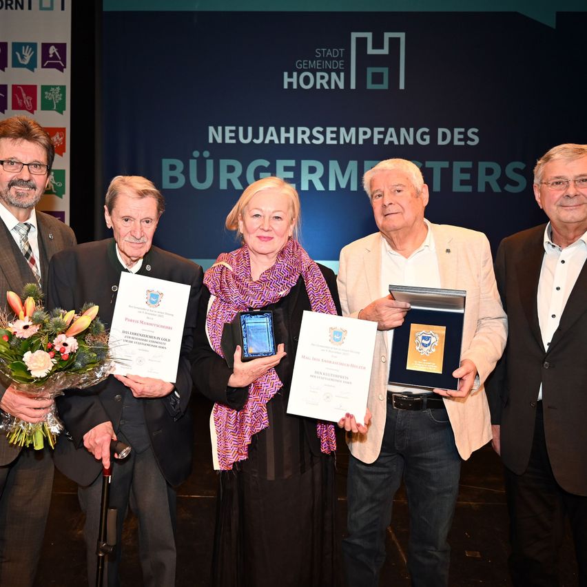 Fünf Personen stehen auf einer Bühne und posieren für ein Foto. Sie halten Urkunden und Auszeichnungen in den Händen. Im Hintergrund steht ein blauer Schriftzug 'Neujahrsempfang des Bürgermeisters'.