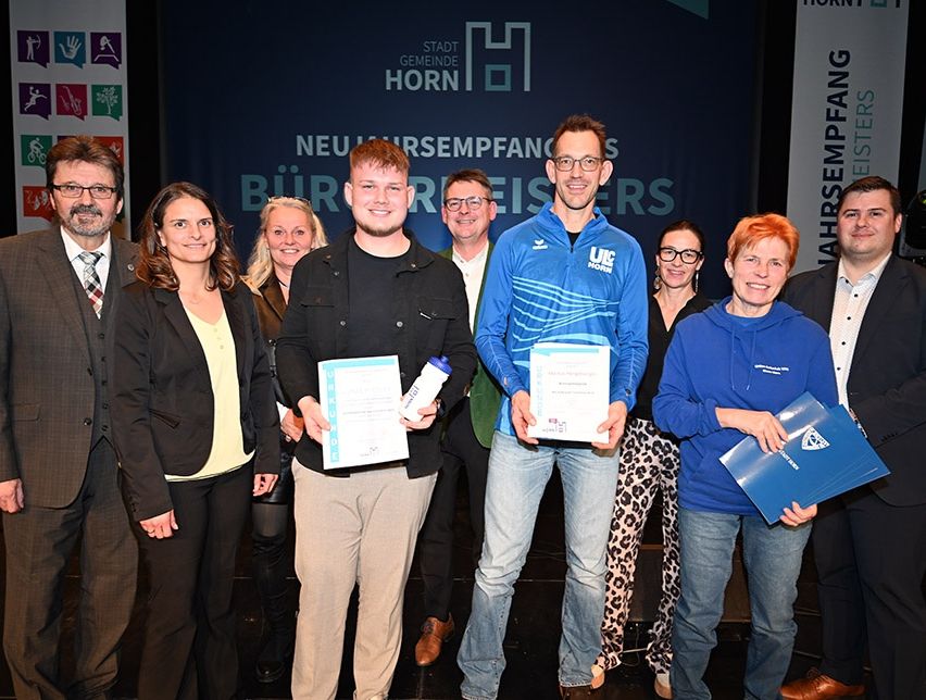 Eine Gruppe von Menschen steht zusammen auf einer Bühne, einige halten Urkunden in den Händen. Sie sind geschäftlich gekleidet, der Mann in der Mitte trägt eine Brille und eine blaue Jacke. Hinter ihnen zeigt ein blauer Hintergrund den Text 'Neuer Gemeindeempfang'.