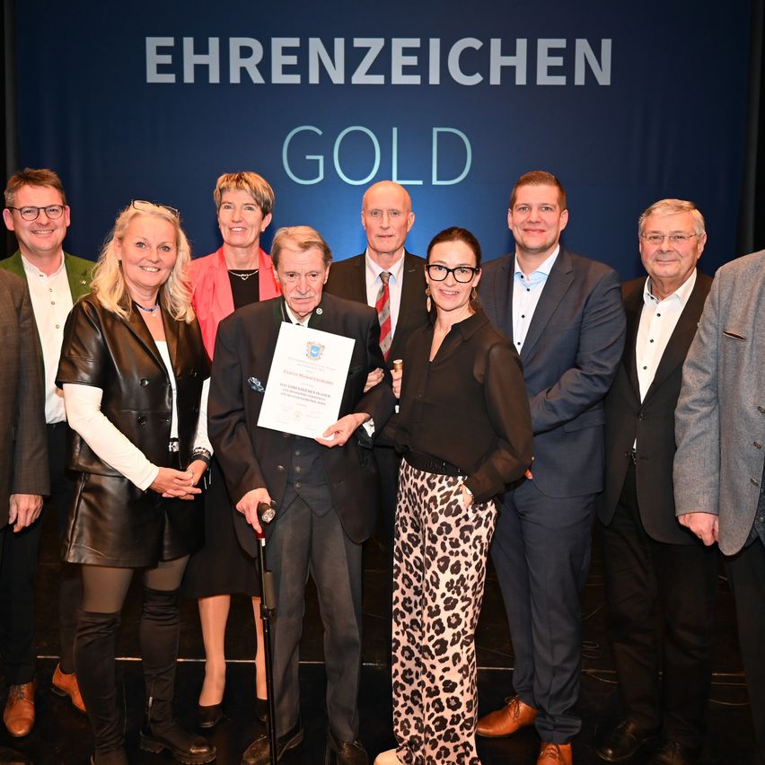 Eine Gruppe von Menschen auf der Bühne, darunter ein Mann mit einem Stock, der ein Zertifikat in der Hand hält, steht vor einem blauen Banner mit der Aufschrift 'Ehrenzeichen Gold'. Sie sind formell gekleidet.