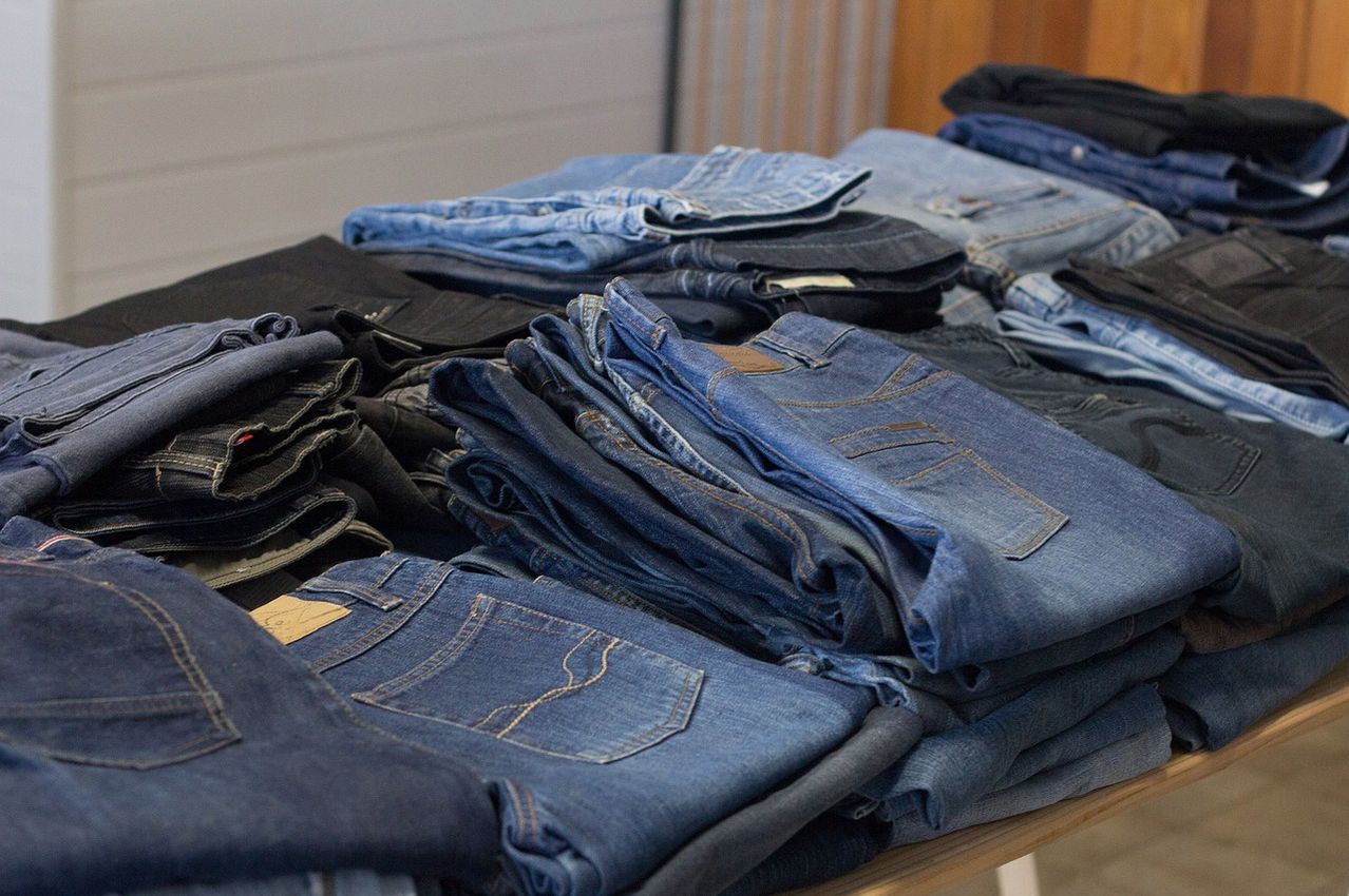 Ein Tisch zeigt eine Sammlung gefalteter Jeans, einschließlich verschiedener Blau- und Schwarztöne. Die Jeans haben unterschiedliche Stile, Taschen und einige Trageetiketten. Der Tisch und der Hintergrund sind schlicht.
