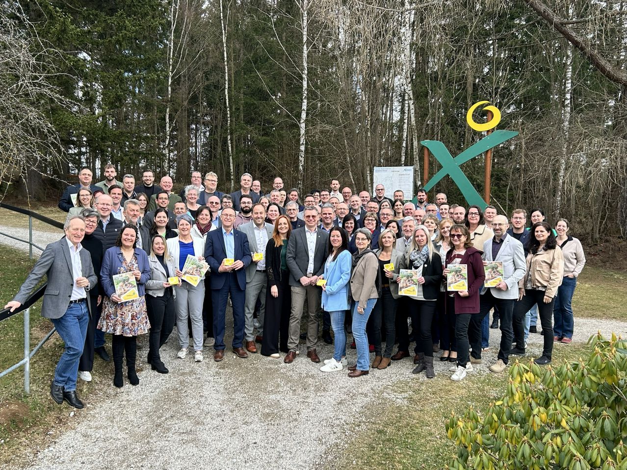 A large group of people stands outdoors, holding pamphlets. They are in front of a green X sign against a backdrop of trees and foliage.