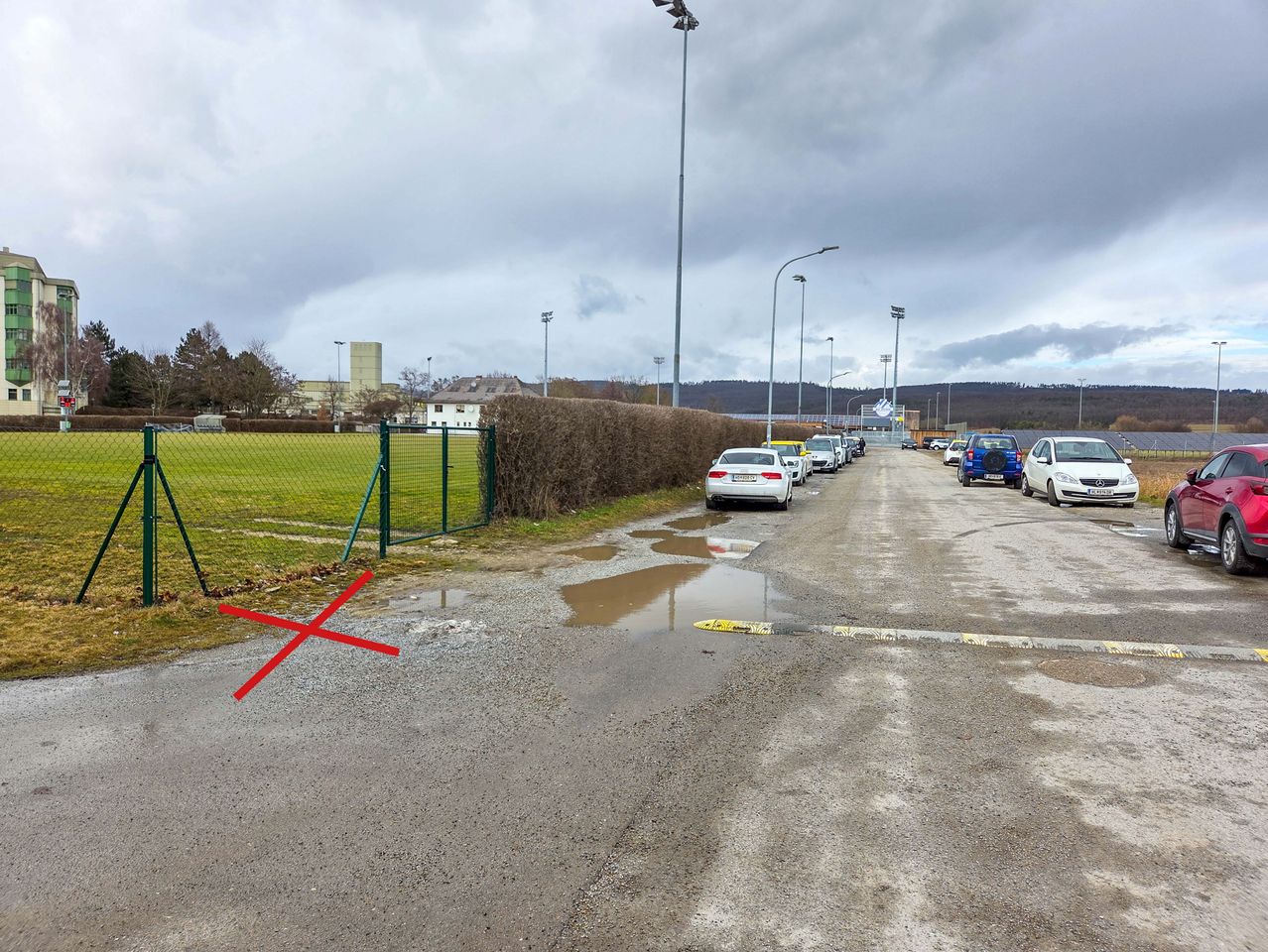 A parking lot with several cars parked, marked with a red cross. The lot has a puddle in the center. A green gate is on the left. Streetlights and trees are in the background.