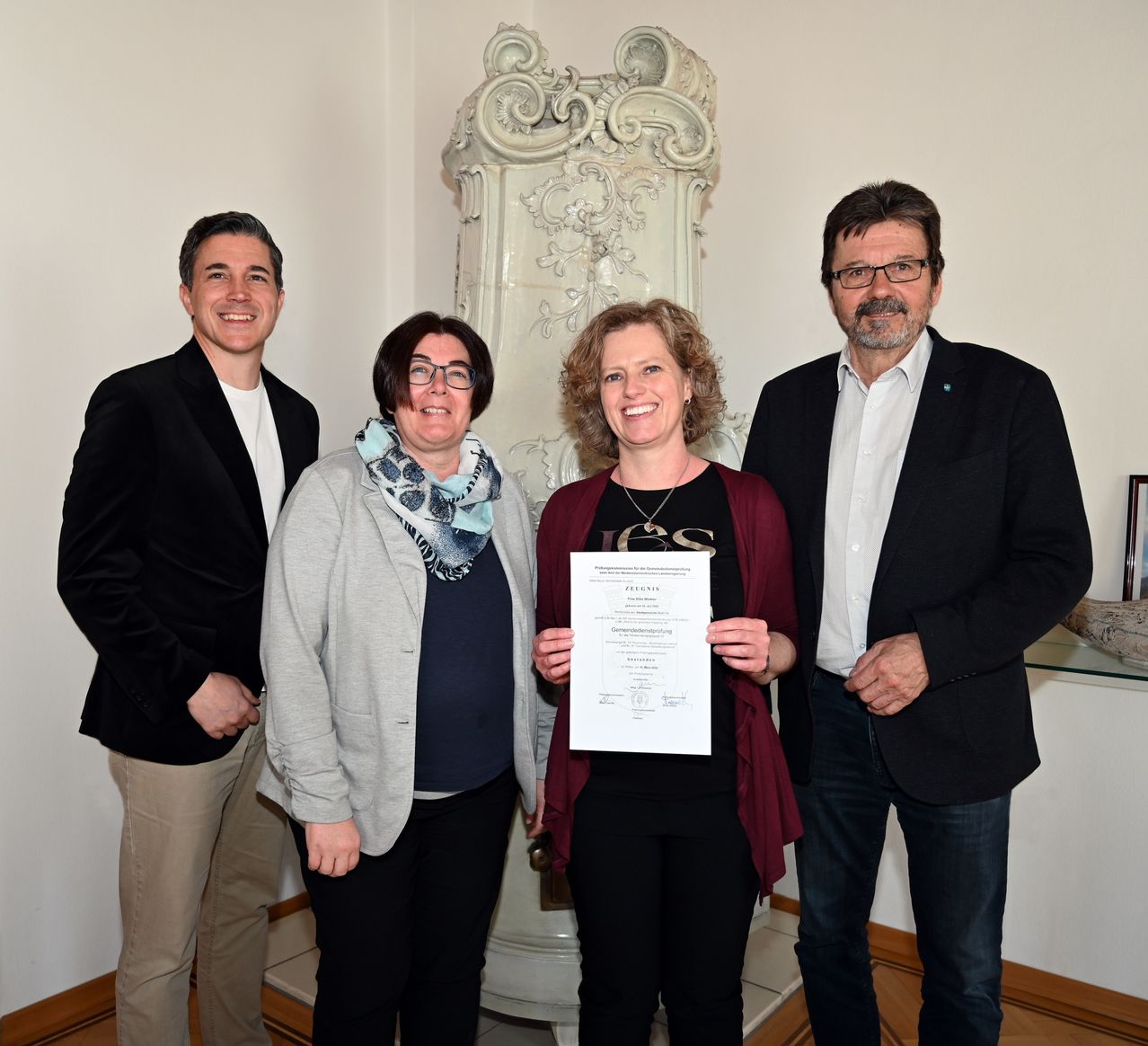 Four individuals stand together, smiling. A woman holds a certificate, while a man on the left smiles. A woman wears glasses and a scarf, and a man on the right wears glasses and a suit. A large white structure is behind them.