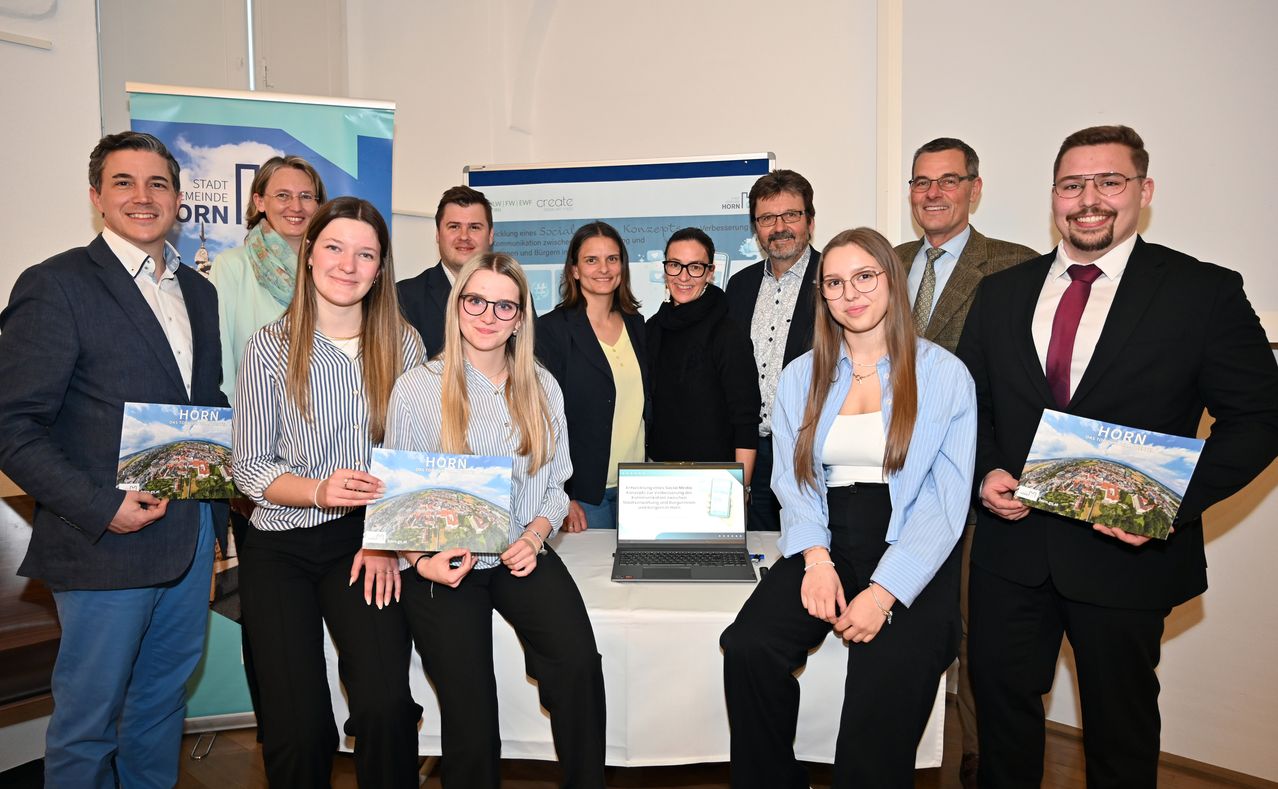 A group of people pose for a photo in front of a desk with a laptop. Some hold books and posters.
