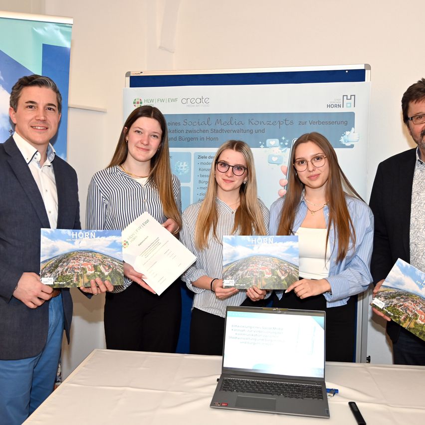 Five individuals stand behind a table with a laptop and certificates in hand. Two of the individuals are holding certificates in front of their chests. Behind them, a large poster with text and logos is visible.