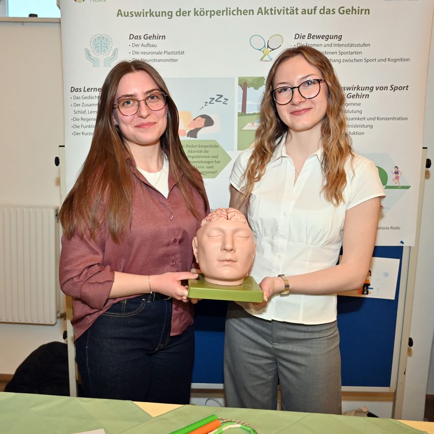 Two women stand before a poster discussing the effects of physical activity on the brain, holding a model of a brain.