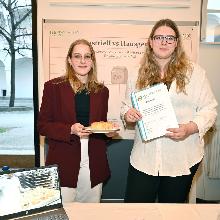 Two women are standing in front of a banner, holding a plate of bread and a certificate. They seem to be posing for a photo. The woman on the right wears glasses and a white blouse. The woman on the left wears a maroon blazer and glasses.