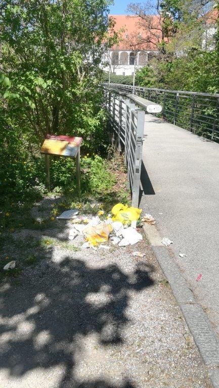 Ein Gehweg mit einem Metallgeländer hat verstreuten Müll und ein kleines Schild auf der Seite in der Nähe von Bäumen und einem Gebäude.