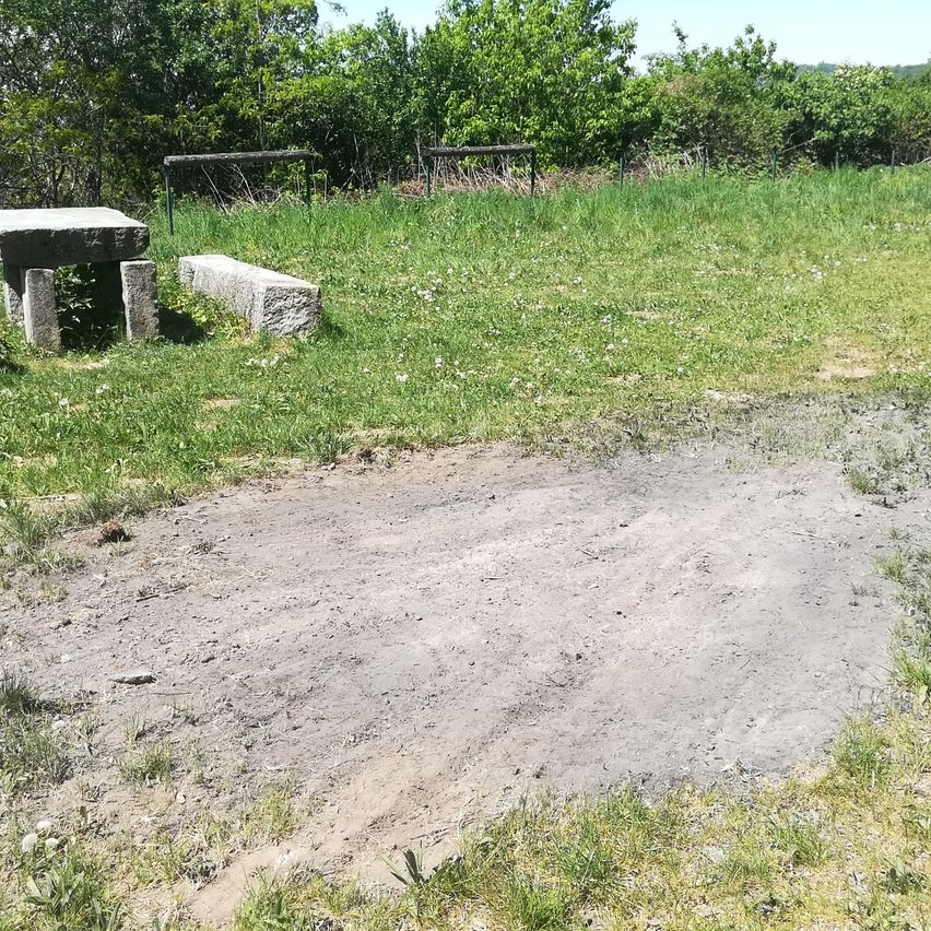 Ein grasiges Feld mit einer Steinbank und einem Steintisch, umgeben von einem Zaun. Der Boden ist aus Erde mit etwas Gras. Hinter der Bank und dem Tisch befinden sich Büsche und Bäume.