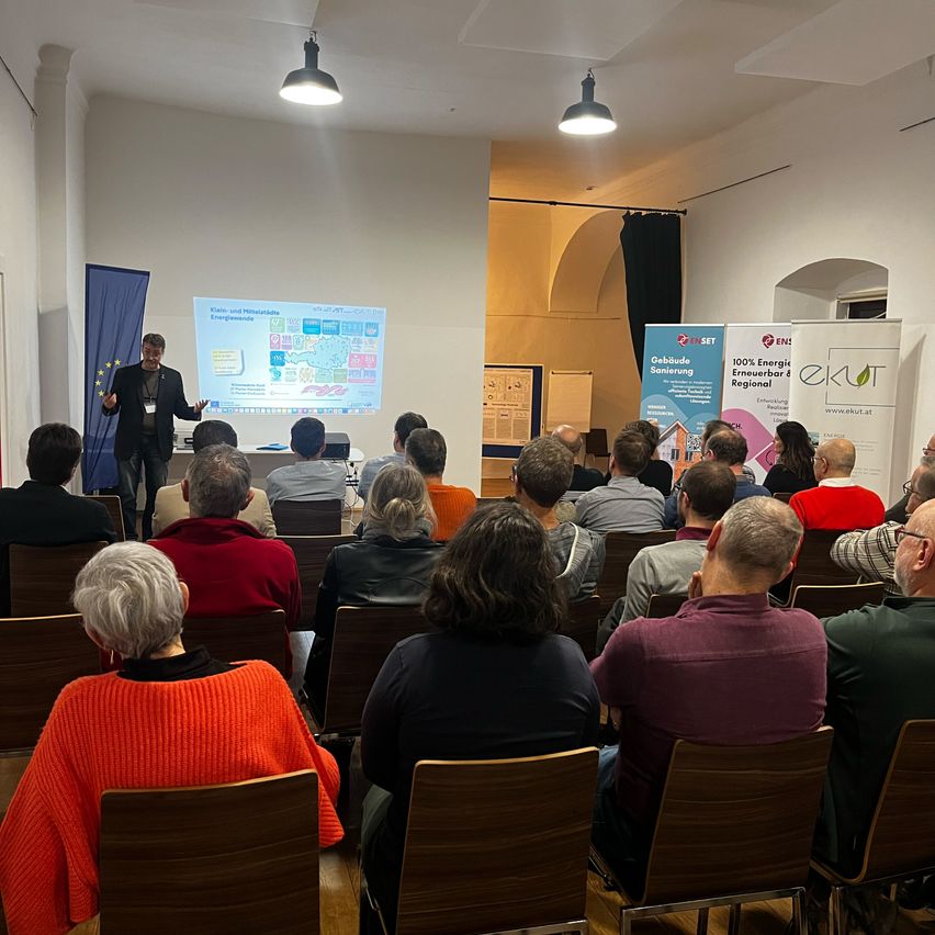 Eine Gruppe von Menschen nimmt in einem Konferenzraum mit Projektorbildschirm, Bannern und Deckenlampen an einer Präsentation teil. Der Moderator steht vorne.