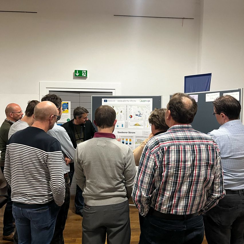 Eine Gruppe von Menschen versammelt sich in einem Raum und betrachtet ein Poster. Es sind hauptsächlich Männer, einige mit Brille. Der Raum hat weiße Wände und einen Holzboden.