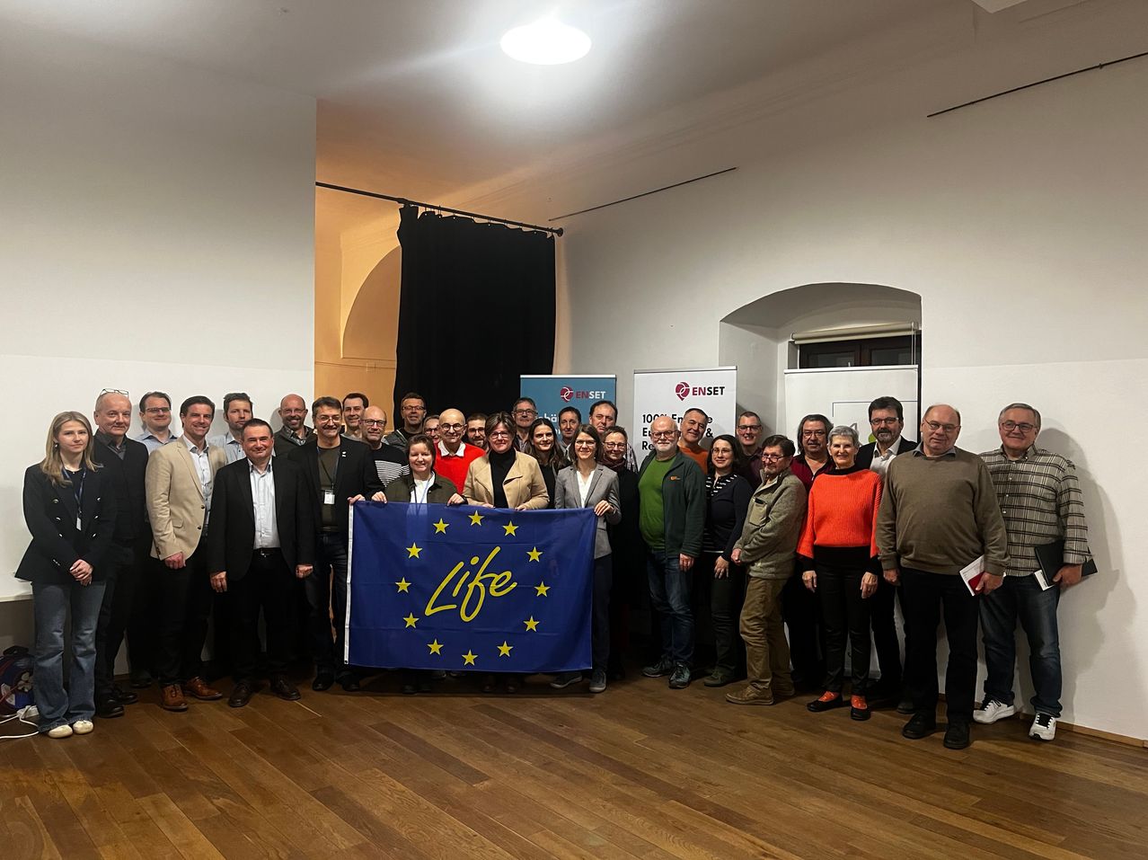 Eine Gruppe von Personen steht in einem Raum zusammen und posiert für ein Foto mit einer Flagge der Europäischen Union, auf der 'Life' steht. Hinter ihnen sind Banner und ein Fenster zu sehen.