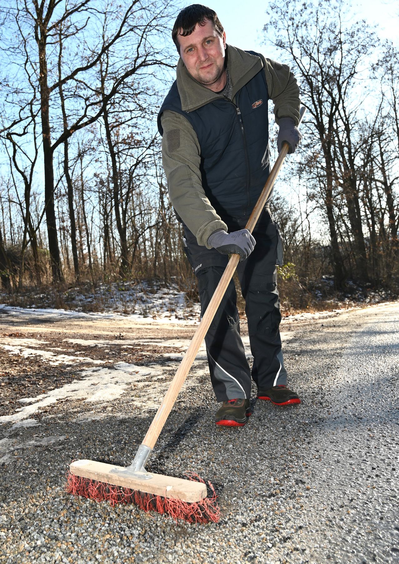 Ein Mann in einer grünen Jacke und Handschuhen fegt die Straße mit einem Besen. Schnee liegt auf dem Boden.