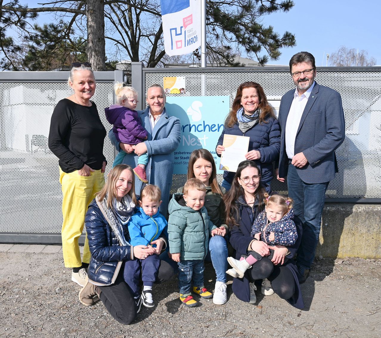 Eine Gruppe von Menschen mit Kindern posiert lächelnd für ein Foto vor einem Zaun. Dahinter zeigt ein Banner Text.