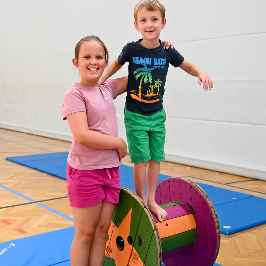 Zwei Kinder stehen auf einer bunten Spule in einer Turnhalle und lächeln für ein Foto. Das Mädchen trägt Rosa, und der Junge trägt ein schwarzes T-Shirt mit einem Stranddesign.