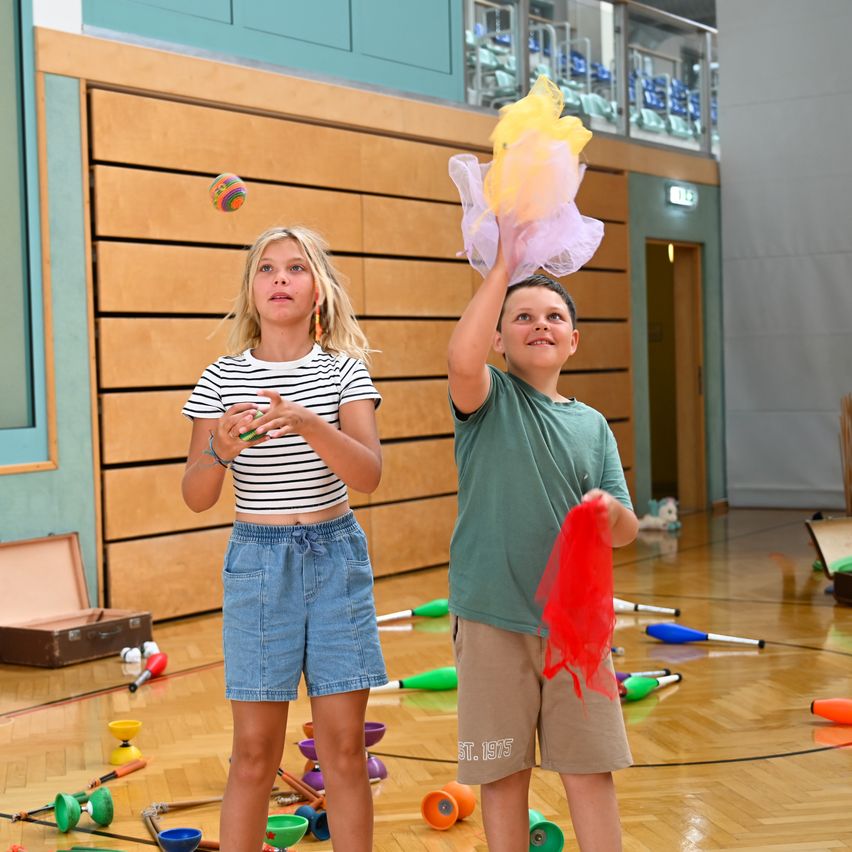 Zwei Kinder spielen mit Jonglierbällen und Tüchern in einer Turnhalle. Das Mädchen hält einen bunten Ball und der Junge ein gelbes und rosafarbenes Tuch.