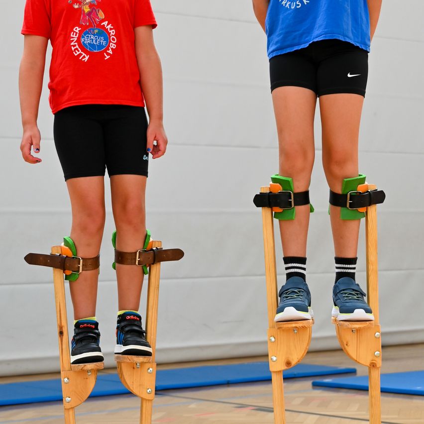 Zwei Kinder stehen auf hölzernen Stelzen, eines trägt ein rotes Shirt und das andere ein blaues. Beide haben Sicherheitsgurte um die Knie. Sie befinden sich in einer Turnhalle mit blauen Matten auf dem Boden.