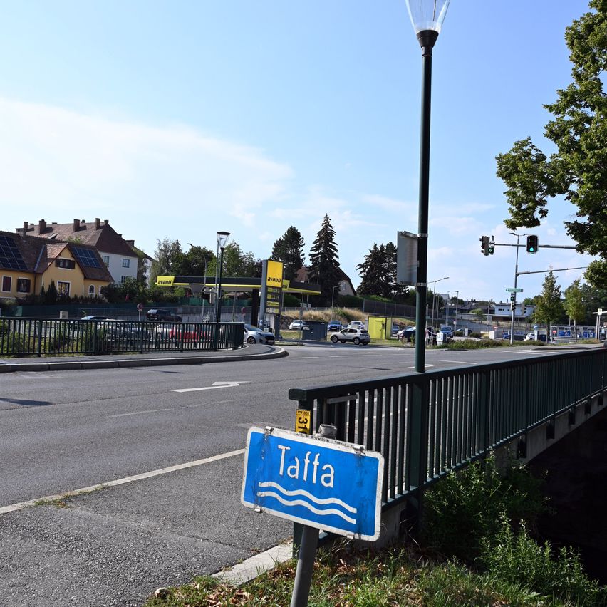 Ein Straßenschild für Taffa steht am Straßenrand, mit einer Brücke und einer Tankstelle im Hintergrund.