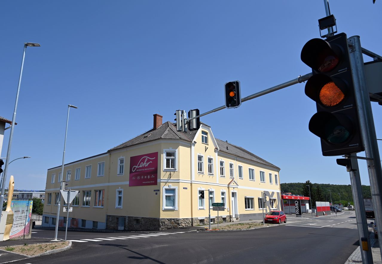 Ein gelbes Gebäude mit einem roten Schild, das 'Lehr' steht, befindet sich an einer Straßenecke, mit einem roten Auto in der Nähe und Ampeln darüber.